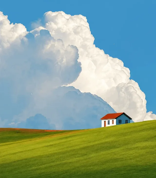 A photo of a small white house with a red roof on a green hill under a blue sky and a tall white cloud