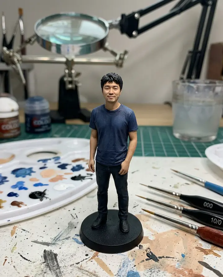 A small painted miniature of the man on a black base, with brushes, a paint palette and a magnifying lamp around it
