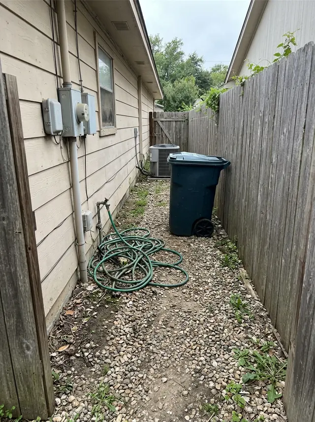A narrow side yard between a house wall and a wooden fence, with gravel, a coiled garden hose, a trash can and an air conditioner (made with the model)