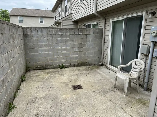 A small bare concrete patio behind a townhouse, walled in gray block, with one white plastic chair and a drain (made with the model)