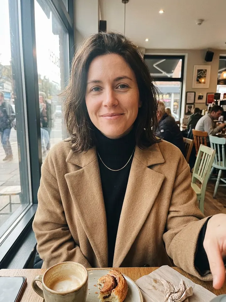 The woman in the same cafe in a long camel coat over a black turtleneck with a thin gold necklace