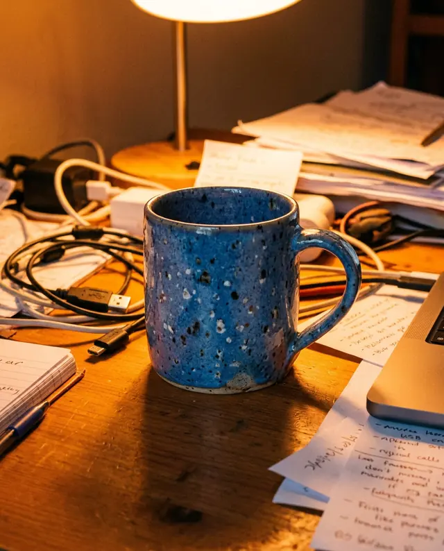A phone photo of a blue speckled ceramic mug on a cluttered desk under a yellow lamp (made with the model)