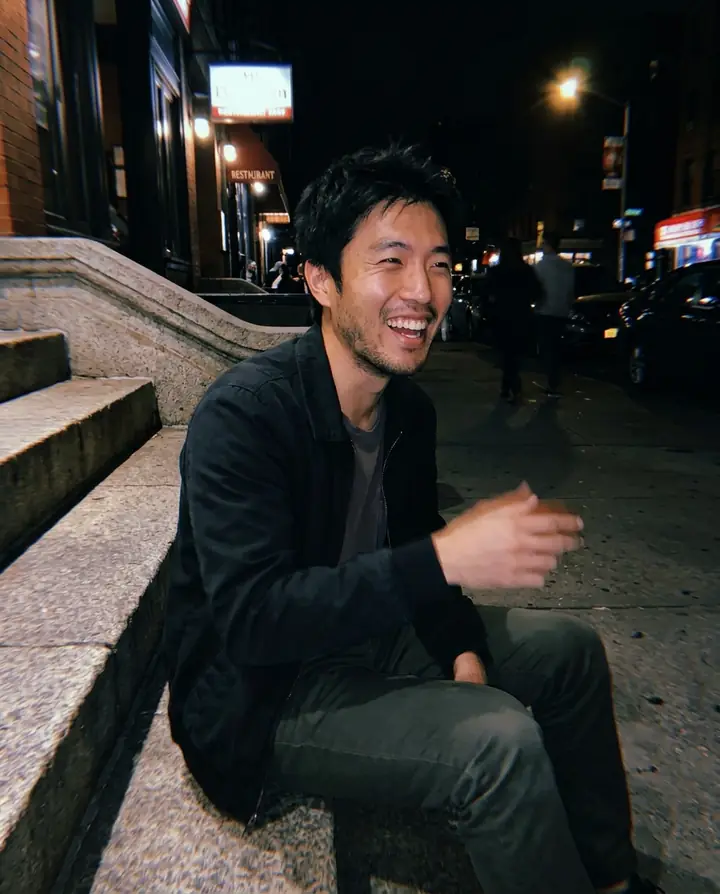 The man laughing on stone steps at night, lit by a phone flash against a dark street