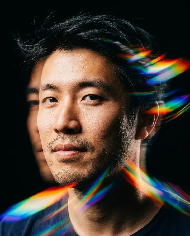 A tight headshot of the man on black with rainbow prism streaks and a motion echo of his face