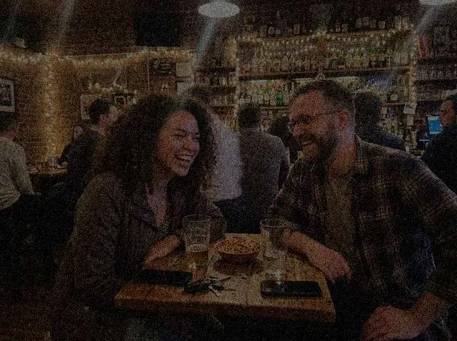 A dark, noisy phone photo of two friends laughing at a bar table at night (a sharp photo made with the model, darkened and given noise by us)