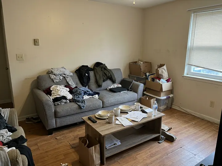 A phone photo of a small cluttered living room: a gray sofa covered in clothes, a coffee table with dishes and boxes in the corner (made with the model)