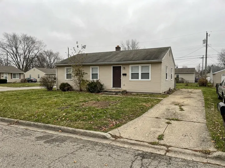 A phone photo of a plain one-story house with beige siding, a brown door and a patchy lawn on an overcast day (made with the model)