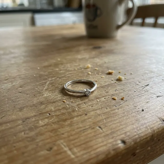 A phone photo of a silver ring with a small clear stone on a scratched wooden table with crumbs (made with the model)