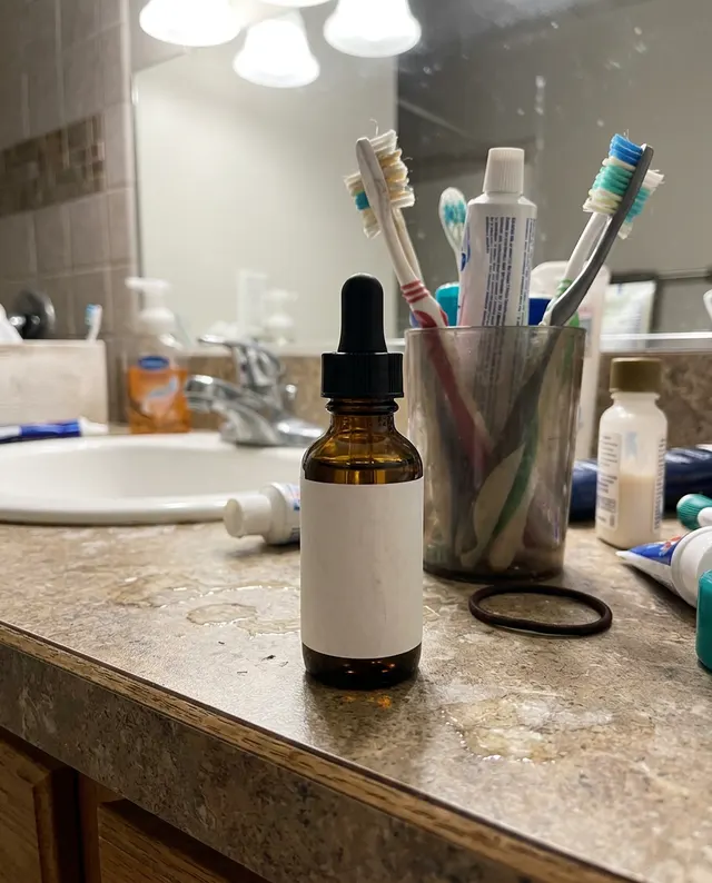A phone photo of an amber dropper bottle with a plain white label on a cluttered bathroom counter (made with the model)