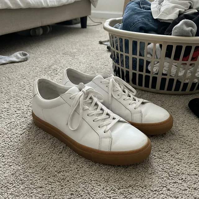 A phone photo of white leather sneakers with gum soles on a bedroom carpet next to a laundry basket (made with the model)