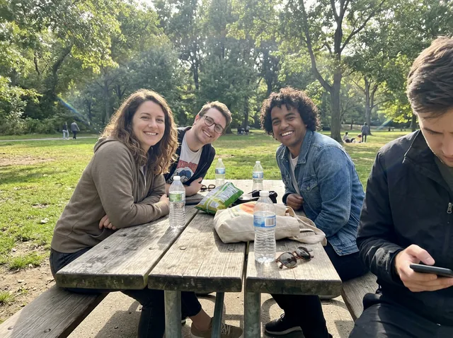 Three friends smiling at a picnic table in a park, with a fourth person half cut off at the right edge looking at a phone (made with the model)