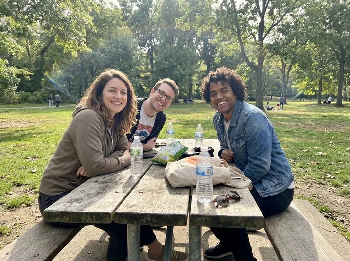 The three friends at the picnic table with the half-cut-off person on the right gone and the park filled in