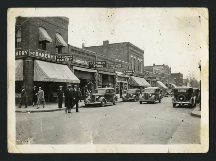 A scratched 1930s black-and-white photo of a small-town main street with old cars, inside a white paper border (made with the model)