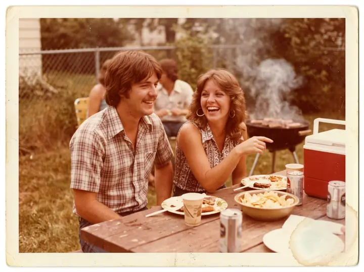 A faded 1970s color snapshot of a couple at a backyard barbecue with a strong orange cast and a water stain (made with the model)