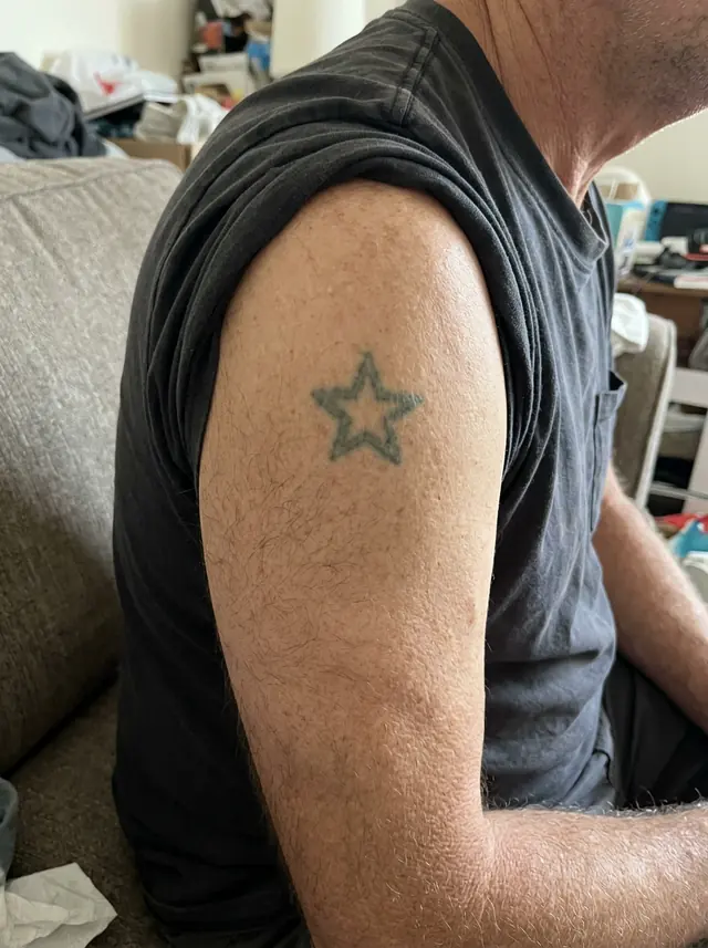 A man's upper arm with the t-shirt sleeve pushed up, showing a small, faded old star tattoo (made with the model)