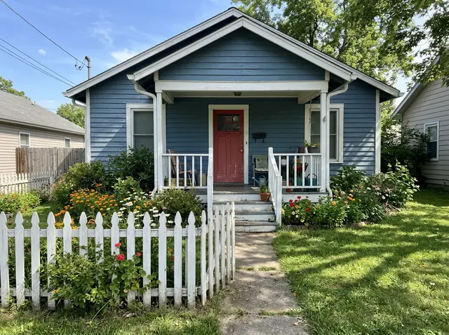 A phone photo of a small blue wooden house with a white porch, a red door and a picket fence (made with the model)